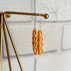 May include: A pair of gold-colored earrings shaped like a baguette. The earrings are hanging from a gold-colored stand.