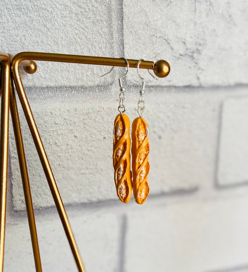 May include: A pair of gold-colored earrings shaped like a baguette. The earrings are hanging from a gold-colored stand.