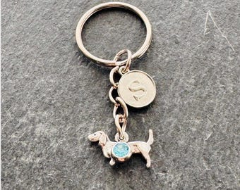 May include: A rose gold-toned metal keychain featuring a circular split ring, a small round charm engraved with the letter S, and a dachshund-shaped charm accented with a single, round, light blue crystal set into the center of the dog's body.