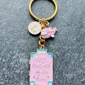 May include: Pink and blue luggage keyring with a gold aeroplane charm and a gold charm with the letter L. The luggage has the text "To travel is to live" on it.