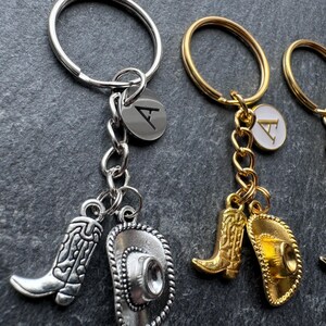 May include: Keyrings with cowboy boot and hat charms. One keyring is silver-toned with a silver boot and hat charm, and a silver initial tag. Two keyrings are gold-toned with gold boot and hat charms, and gold initial tags.