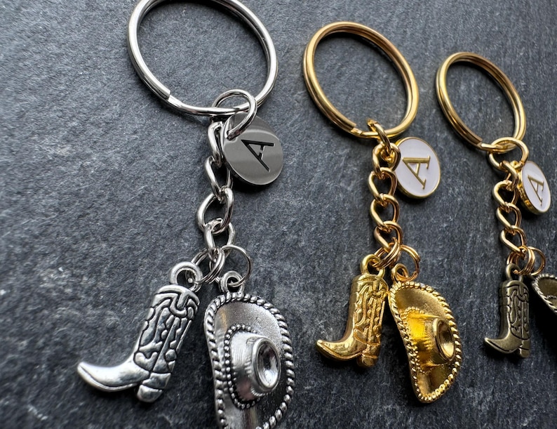 May include: Keyrings with cowboy boot and hat charms. One keyring is silver-toned with a silver boot and hat charm, and a silver initial tag. Two keyrings are gold-toned with gold boot and hat charms, and gold initial tags.