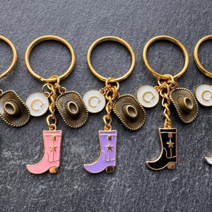 May include: Five keyrings with gold rings, each featuring a cowboy boot charm in red, pink, purple, black, and white. Each keyring also includes a small cowboy hat charm and a white disc with a gold letter "C".