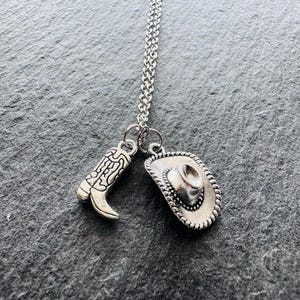 May include: Silver necklace with two charms: a cowboy boot and a cowboy hat. The boot charm has etched details, and the hat charm has a rope-like border. The necklace hangs on a silver chain, ideal for Western-themed jewellery.