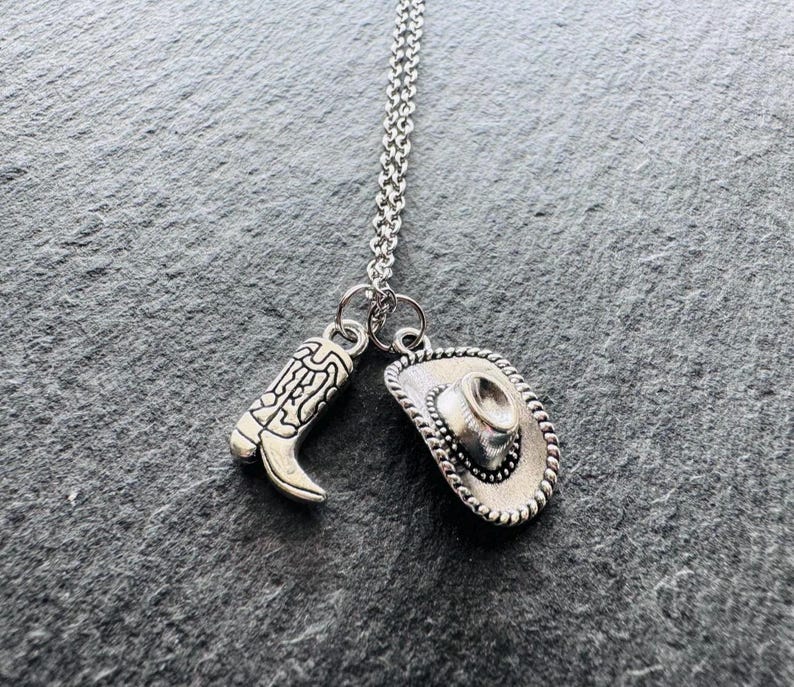 May include: Silver necklace with two charms: a cowboy boot and a cowboy hat. The boot charm has etched details, and the hat charm has a rope-like border. The necklace hangs on a silver chain, ideal for Western-themed jewellery.