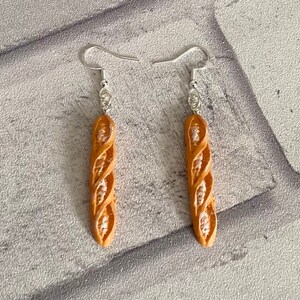 May include: A pair of dangle earrings featuring a brown, twisted, bread-shaped charm. The earrings have silver hooks.