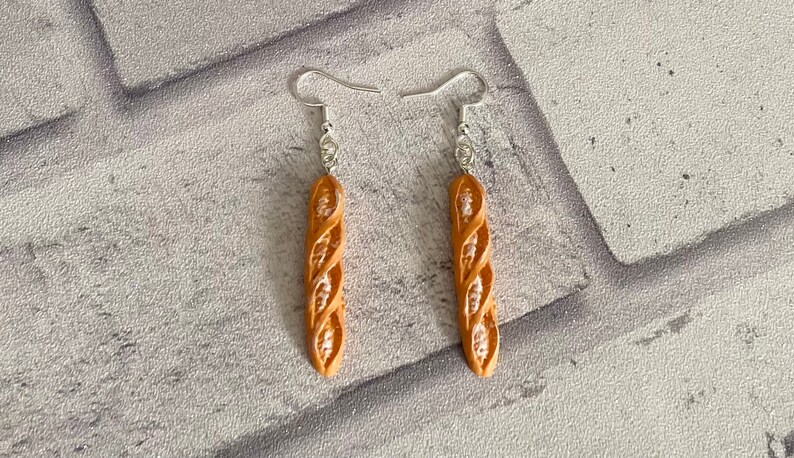 May include: A pair of dangle earrings featuring a brown, twisted, bread-shaped charm. The earrings have silver hooks.