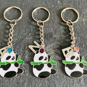 May include: Three silver keychains with panda bear charms. Each charm features a different letter, A, N, and D, and a jeweled stone. The charms are holding a green bamboo shoot.