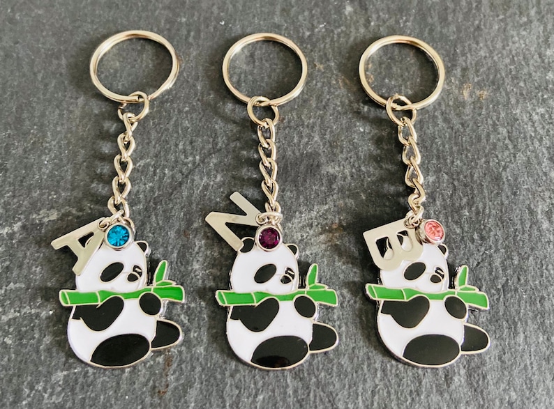 May include: Three silver keychains with panda bear charms. Each charm features a different letter, A, N, and D, and a jeweled stone. The charms are holding a green bamboo shoot.