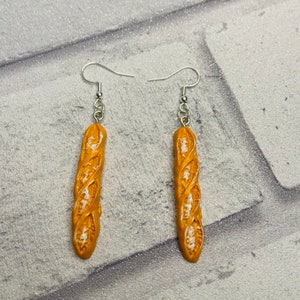 May include: A pair of dangle earrings featuring a realistic-looking baguette bread charm. The charms are a light brown color with white details and are attached to silver hooks.