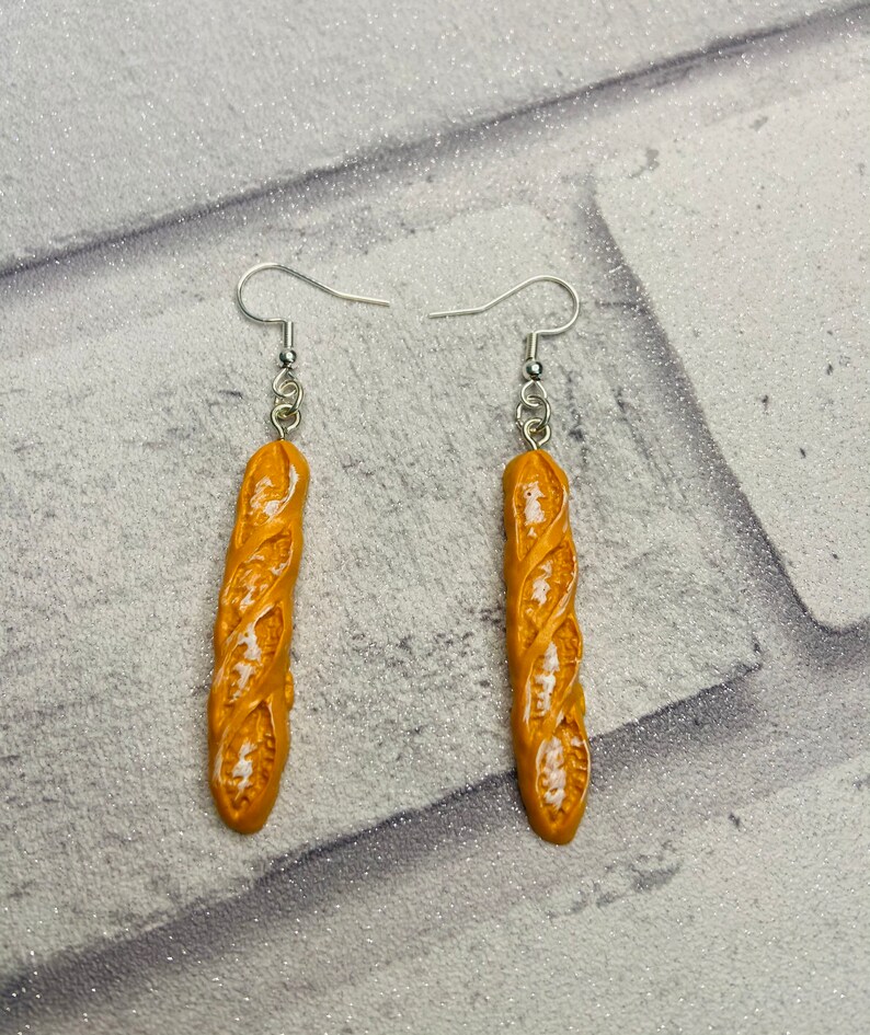 May include: A pair of dangle earrings featuring a realistic-looking baguette bread charm. The charms are a light brown color with white details and are attached to silver hooks.