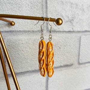 May include: A pair of dangle earrings featuring a realistic-looking baguette bread charm in a light yellow color. The earrings are hanging from silver hooks.