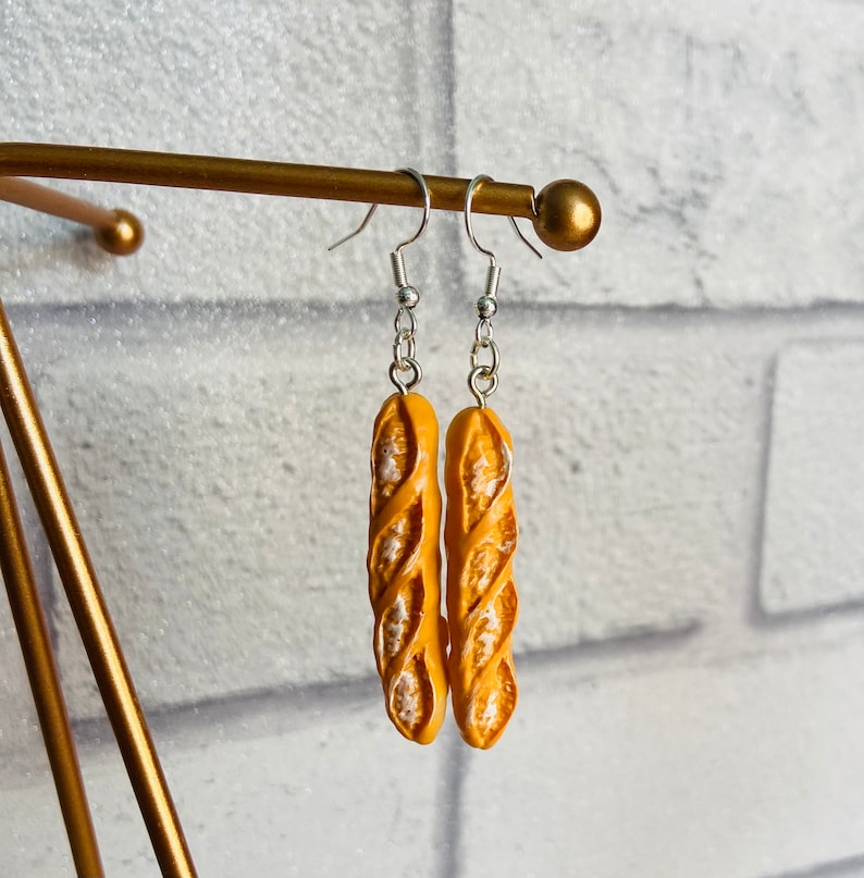 May include: A pair of dangle earrings featuring a realistic-looking baguette bread charm in a light yellow color. The earrings are hanging from silver hooks.