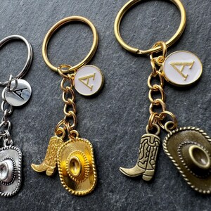 May include: Keyrings with cowboy boot and hat charms. One keyring is silver, and the other two are gold. Each keyring has a round charm with the letter "A". The charms are attached to a keyring with a chain.