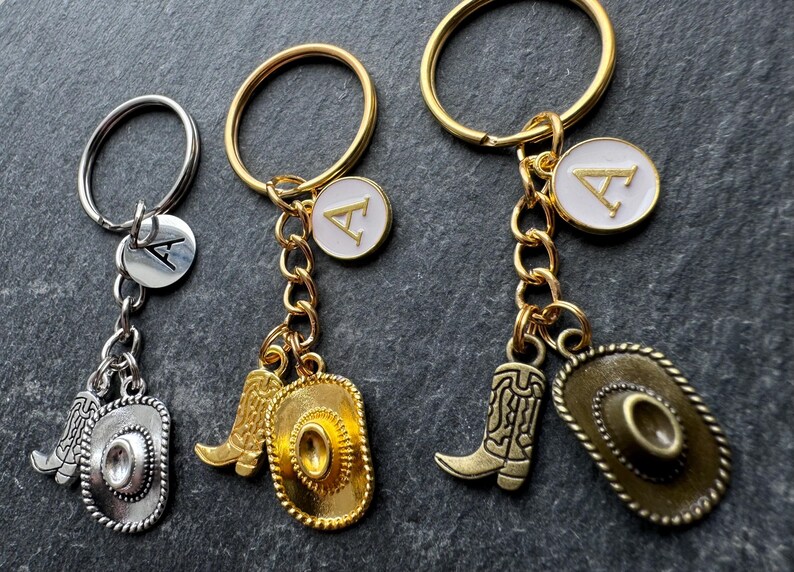 May include: Keyrings with cowboy boot and hat charms. One keyring is silver, and the other two are gold. Each keyring has a round charm with the letter "A". The charms are attached to a keyring with a chain.