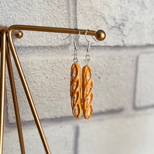 May include: A pair of silver earrings with a baguette-shaped charm. The charms are a light brown color and have a textured surface.