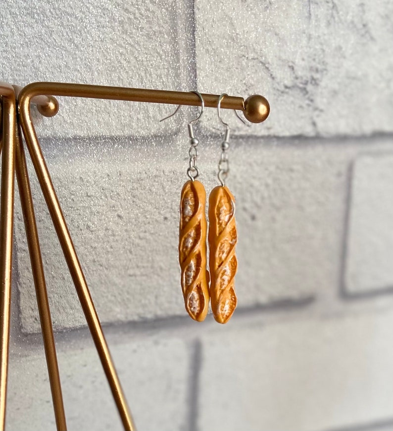 May include: A pair of silver earrings with a baguette-shaped charm. The charms are a light brown color and have a textured surface.