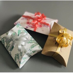 May include: Three pillow boxes with different designs and colors. The first box is white with green leaves and a silver ribbon. The second box is white with pink and red flowers and a red ribbon. The third box is brown with a gold ribbon.