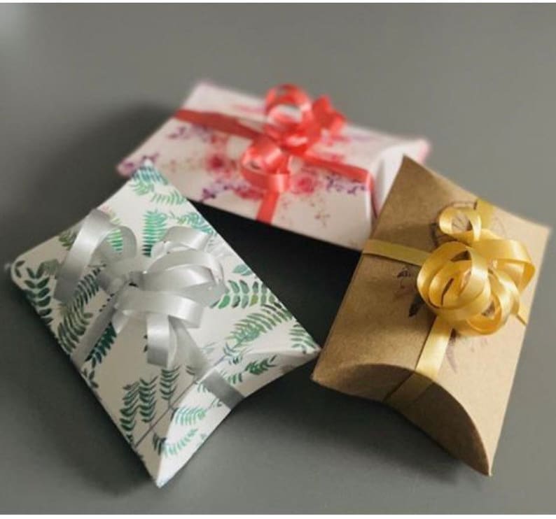 May include: Three pillow boxes with different designs and colors. The first box is white with green leaves and a silver ribbon. The second box is white with pink and red flowers and a red ribbon. The third box is brown with a gold ribbon.