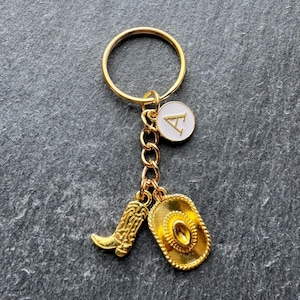 May include: Gold-coloured keyring with a cowboy theme. The keyring features a gold ring, a chain, and three charms: a cowboy boot, a cowboy hat, and a white circular charm with the letter "A".