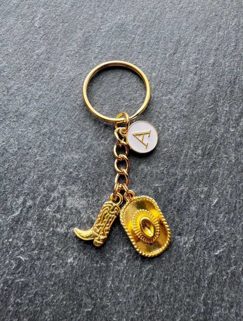 May include: Gold-coloured keyring with a cowboy theme. The keyring features a gold ring, a chain, and three charms: a cowboy boot, a cowboy hat, and a white circular charm with the letter "A".