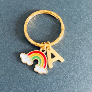 May include: Gold keyring with a rainbow and the letter A charm. The rainbow is red, yellow, green, and blue with white clouds.