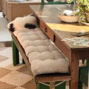 May include: A rustic wooden dining table with a green bench and a striped cushion. A fluffy, black and white pillow rests on the bench. The table has a drawer and is set with cards and a basket.