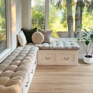 May include: An L-shaped white wooden window bench with built-in storage drawers and metal pulls. The bench features thick, tufted beige cushions and is decorated with a collection of square and round throw pillows in white, olive, and patterned fabric.