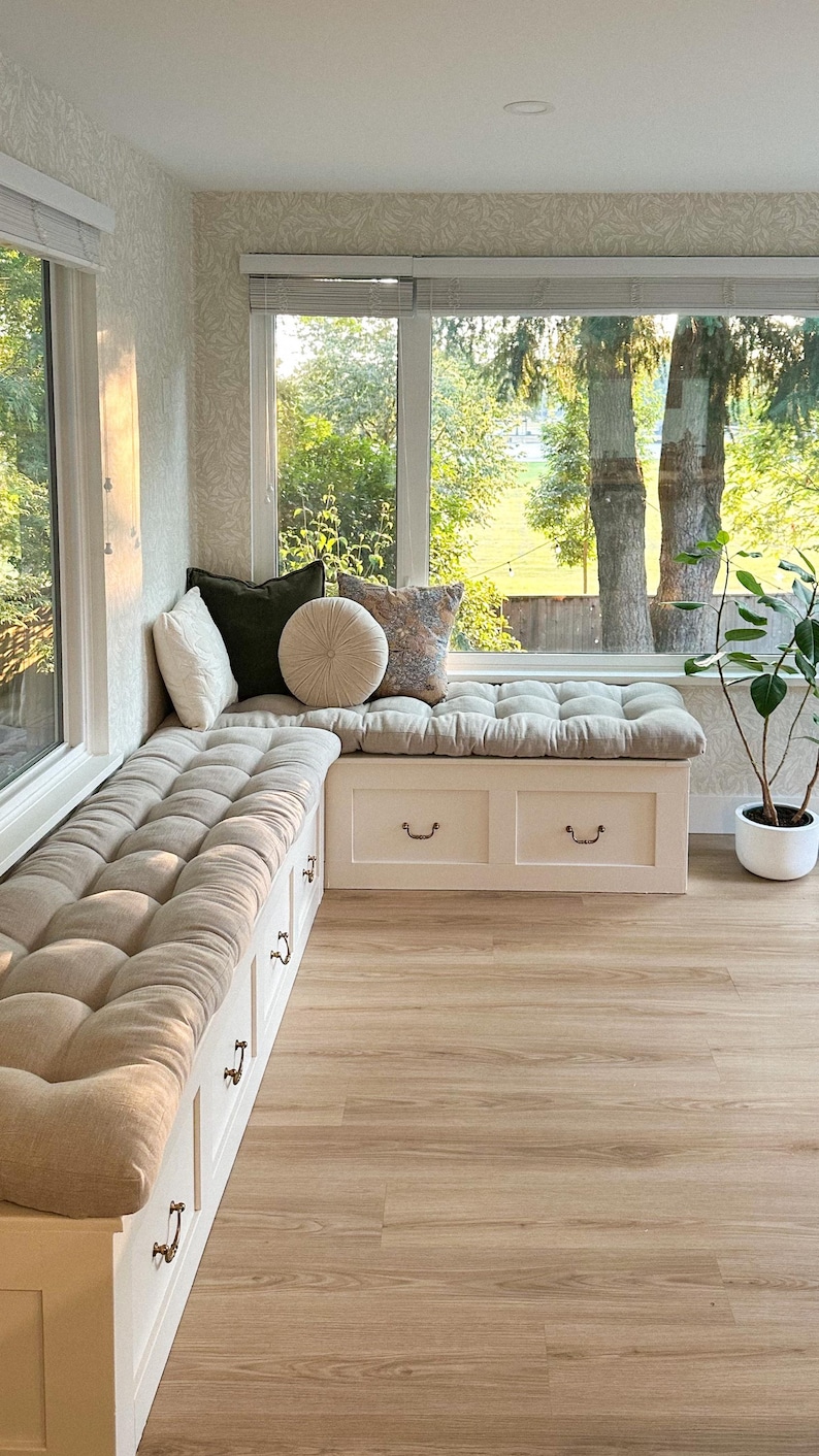 May include: An L-shaped white wooden window bench with built-in storage drawers and metal pulls. The bench features thick, tufted beige cushions and is decorated with a collection of square and round throw pillows in white, olive, and patterned fabric.