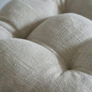 May include: A round, tufted seat cushion featuring a light beige, textured fabric with a visible woven grain. The cushion has deep indentations created by central tufting points, giving the surface a soft, pillowed, and dimensional appearance.