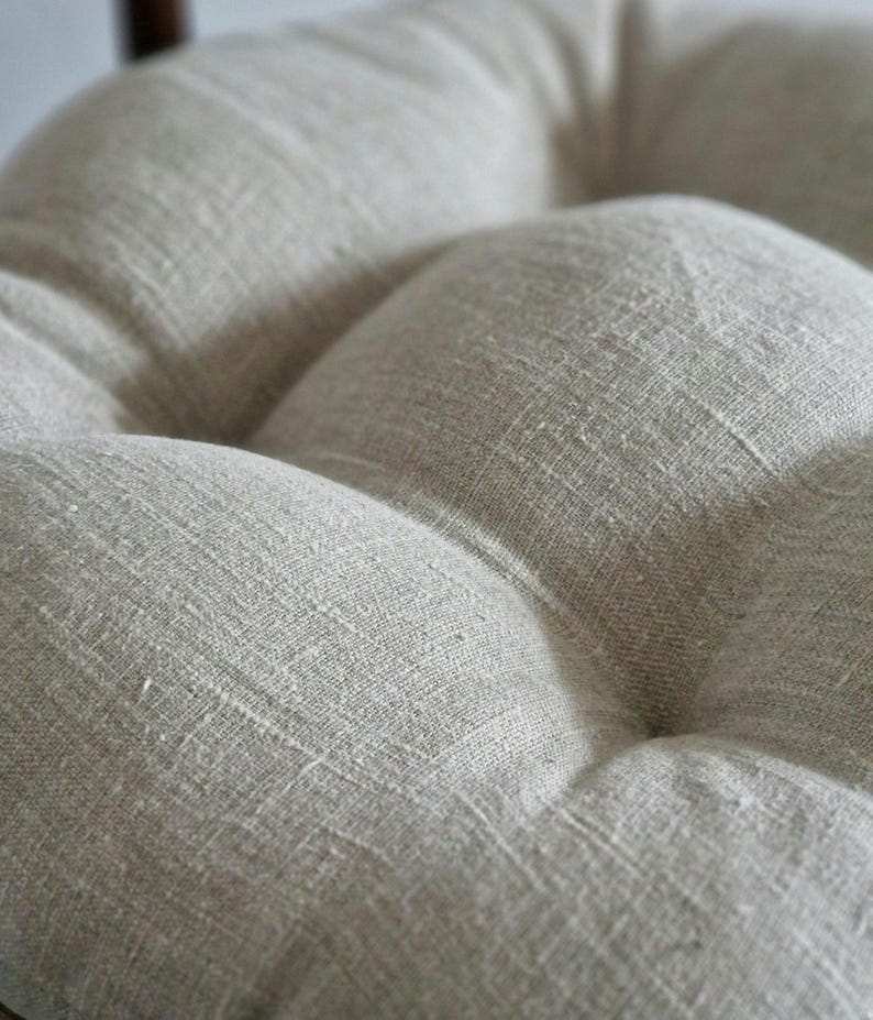 May include: A round, tufted seat cushion featuring a light beige, textured fabric with a visible woven grain. The cushion has deep indentations created by central tufting points, giving the surface a soft, pillowed, and dimensional appearance.