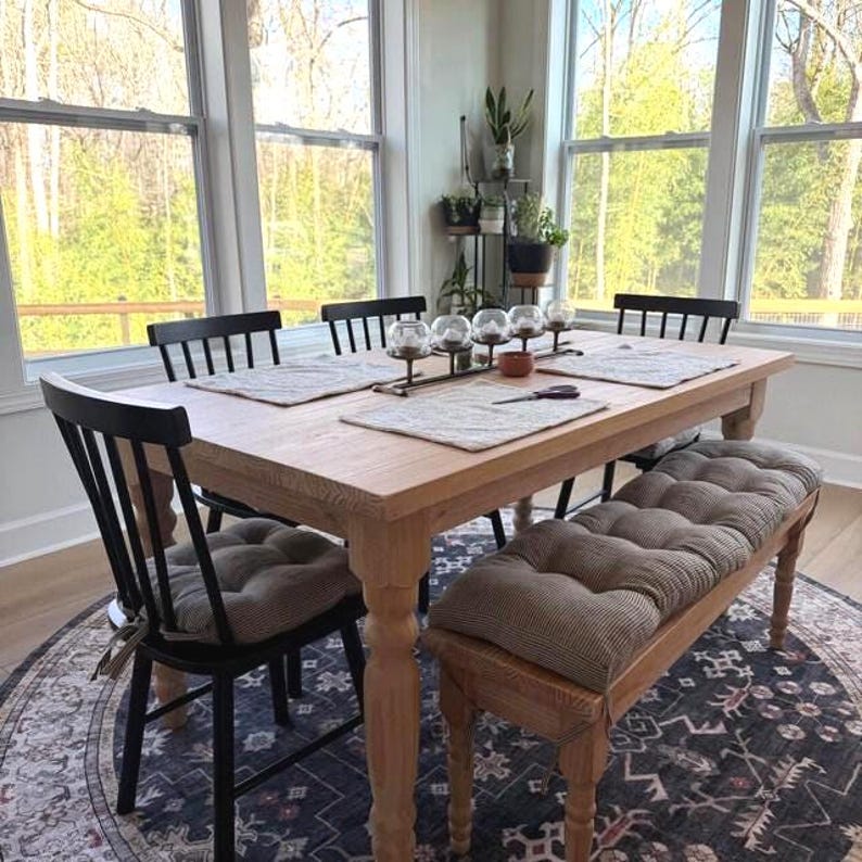 May include: A light-toned wooden dining table with four black spindle-back chairs and a matching wooden bench. The table features a central metal candle holder with five glass globes, light-colored placemats, and tufted brown fabric seat cushions.