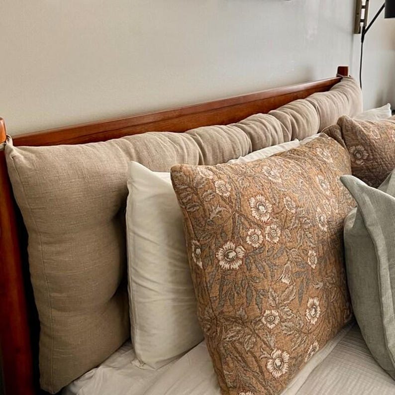 May include: A wooden headboard featuring a long, tufted beige linen cushion attached to the frame. In front, a white pillow sits beside a rectangular pillow with a brown and white floral pattern, and a light green pillow is partially visible.