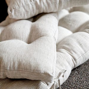 May include: A tufted bench cushion featuring a beige and white vertical pinstripe pattern. The cushion is constructed with multiple stitched sections to create a plush, padded appearance and includes fabric ties at the corner for secure attachment.