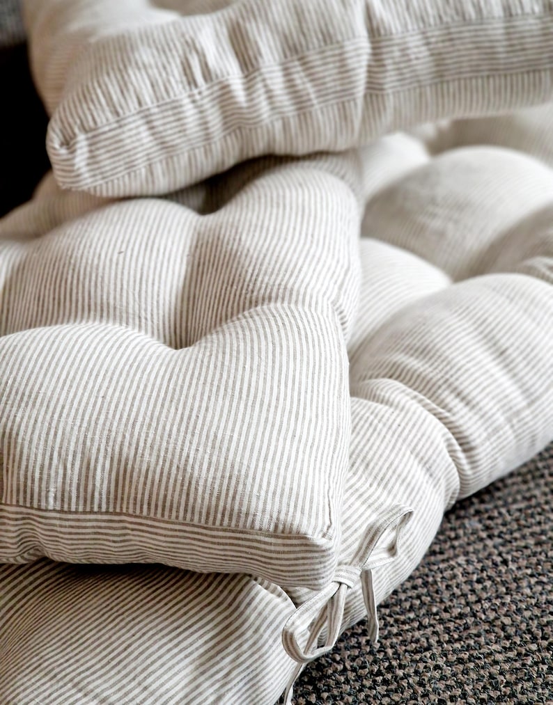 May include: A tufted bench cushion featuring a beige and white vertical pinstripe pattern. The cushion is constructed with multiple stitched sections to create a plush, padded appearance and includes fabric ties at the corner for secure attachment.