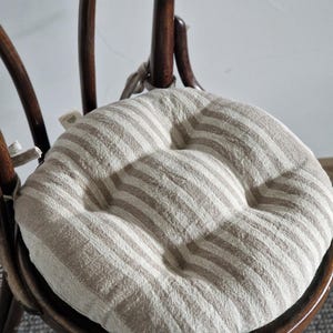 May include: A round, beige and white striped linen chair cushion with a tufted centre. The cushion is placed on a wooden chair.
