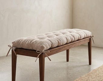 May include: A rectangular wooden bench with four straight legs, topped with a thick, tufted cushion featuring a beige and white pinstripe pattern. The cushion is secured to the bench frame with fabric ties at each of the four corners.
