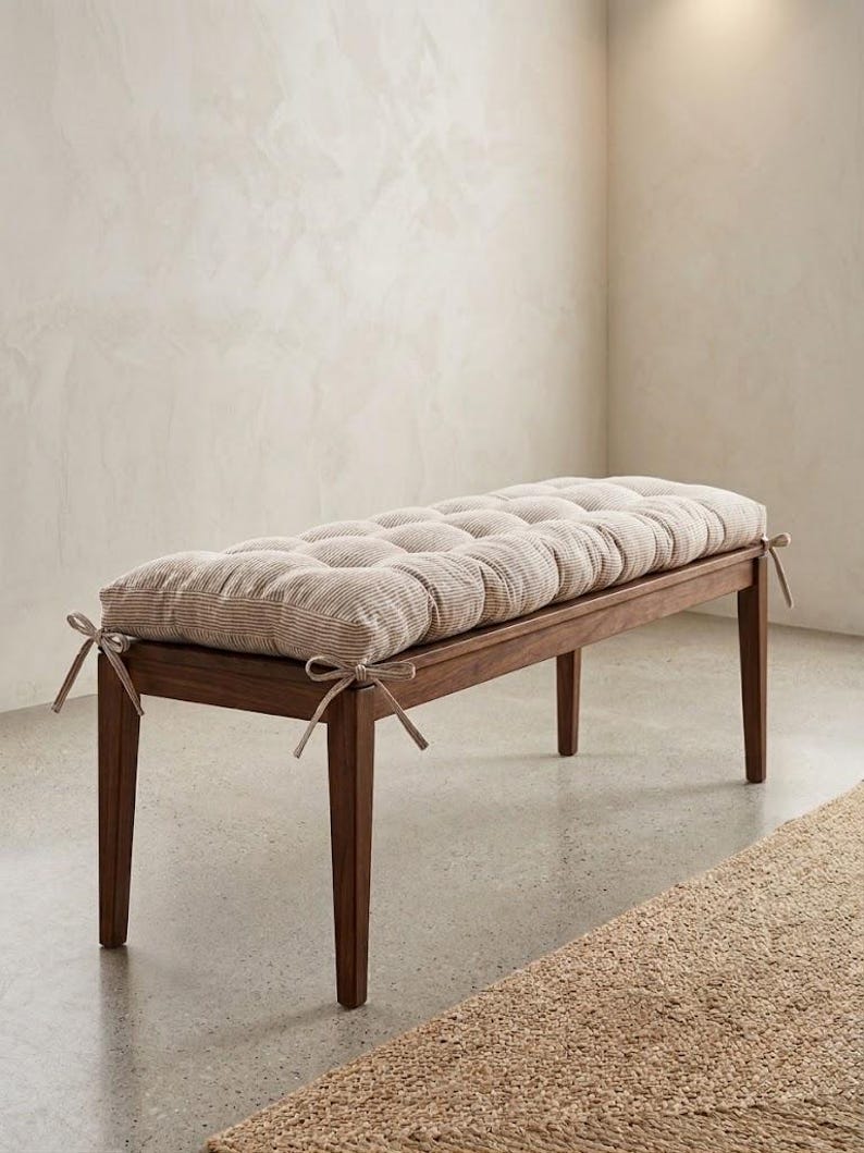 May include: A rectangular wooden bench with four straight legs, topped with a thick, tufted cushion featuring a beige and white pinstripe pattern. The cushion is secured to the bench frame with fabric ties at each of the four corners.