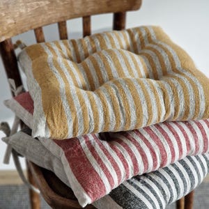 Puede incluir: Tres cojines de silla a rayas apilados en una silla de madera. El cojín superior es amarillo y gris, el cojín del medio es rojo y blanco, y el cojín inferior es negro y blanco.