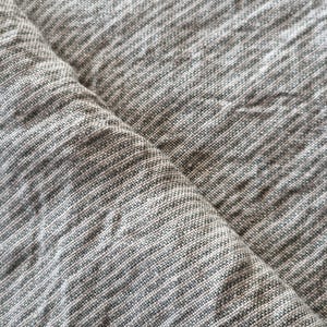 May include: A piece of fabric featuring a heathered gray and white color palette with a visible woven texture. The material displays a subtle cross-hatch pattern created by the interlacing of light and dark threads, resulting in a soft, mottled appearance across the surface of the textile.