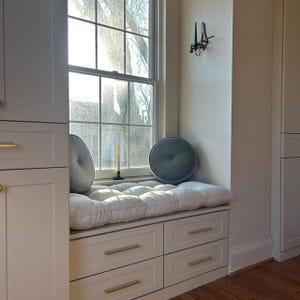 May include: A window seat featuring a long, tufted, off-white cushion with thin grey pinstripes, two round grey velvet pillows, and four drawers with gold-toned handles. A tall, thin candle sits on the windowsill near a wall-mounted candle holder.