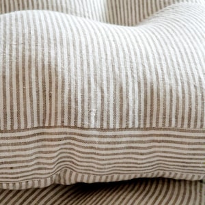May include: A tufted cushion featuring a woven fabric with a pattern of thin, vertical beige and white stripes. The cushion has a soft, plush appearance with visible indentations created by the tufting, showcasing a textured, natural textile finish.