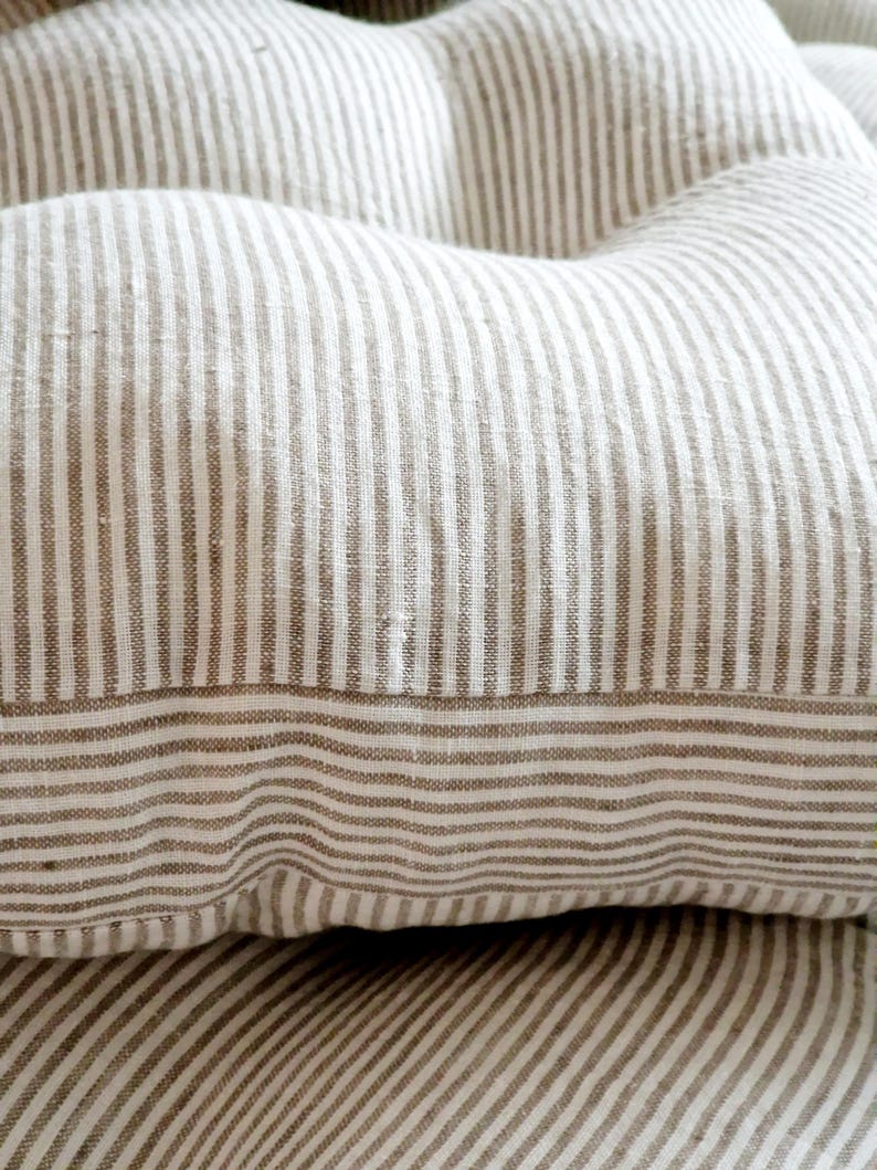 May include: A tufted cushion featuring a woven fabric with a pattern of thin, vertical beige and white stripes. The cushion has a soft, plush appearance with visible indentations created by the tufting, showcasing a textured, natural textile finish.