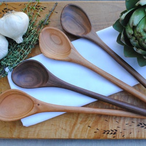 May include: Four wooden spoons in shades of brown are arranged on a wooden chopping board with a white cloth. Two garlic bulbs, thyme sprigs, and an artichoke are also present. The spoons are for kitchen use.