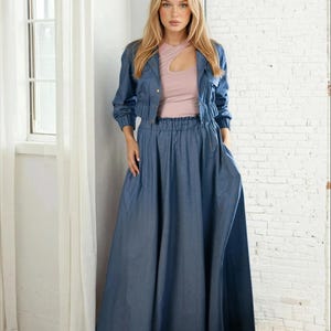 May include: A woman wearing a denim jacket, a light pink top, and a long denim skirt. The jacket is cropped and buttoned, and the skirt is a flowing, full length style. The outfit is a casual, yet stylish ensemble.