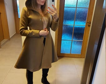 May include: A tan hooded coat featuring a flared silhouette, long sleeves, and a front button closure. The garment is worn over a light-colored top, black trousers, and black boots, creating a structured look with a pointed hood design.