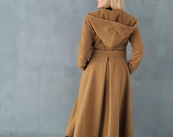 Puede incluir: Un abrigo largo de color camel con capucha, mangas largas y cinturón en la cintura. El abrigo presenta una falda acampanada hasta el suelo con un pliegue central en la espalda y bolsillos laterales, combinado con botas y leggings negros.