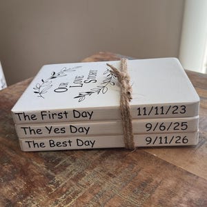 May include: A stack of three white, rectangular blocks tied with twine. Each block is labeled with a special day: "The First Day", "The Yes Day", and "The Best Day". Dates are printed on the side: 11/11/23, 9/6/25, and 9/11/26. The top block has the text "Our Love Story".