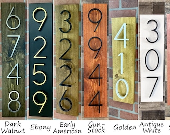 May include: Ten different house numbers, each on a separate wooden sign, are displayed in a vertical orientation. The numbers are made of metal and are painted in various colors, including gold, black, and white. The signs are painted in different wood stains, including dark walnut, ebony, early American, gunstock, golden, antique white, Sedona, and worn navy.