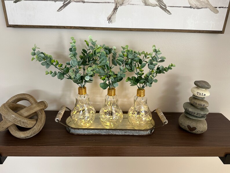 May include: Three clear glass light bulb vases with gold tops, filled with green eucalyptus sprigs, are arranged in a metal tray. The tray is on a dark brown shelf with a wooden knot and a stack of gray stones with the words "This is our Happy Place" inscribed on them.